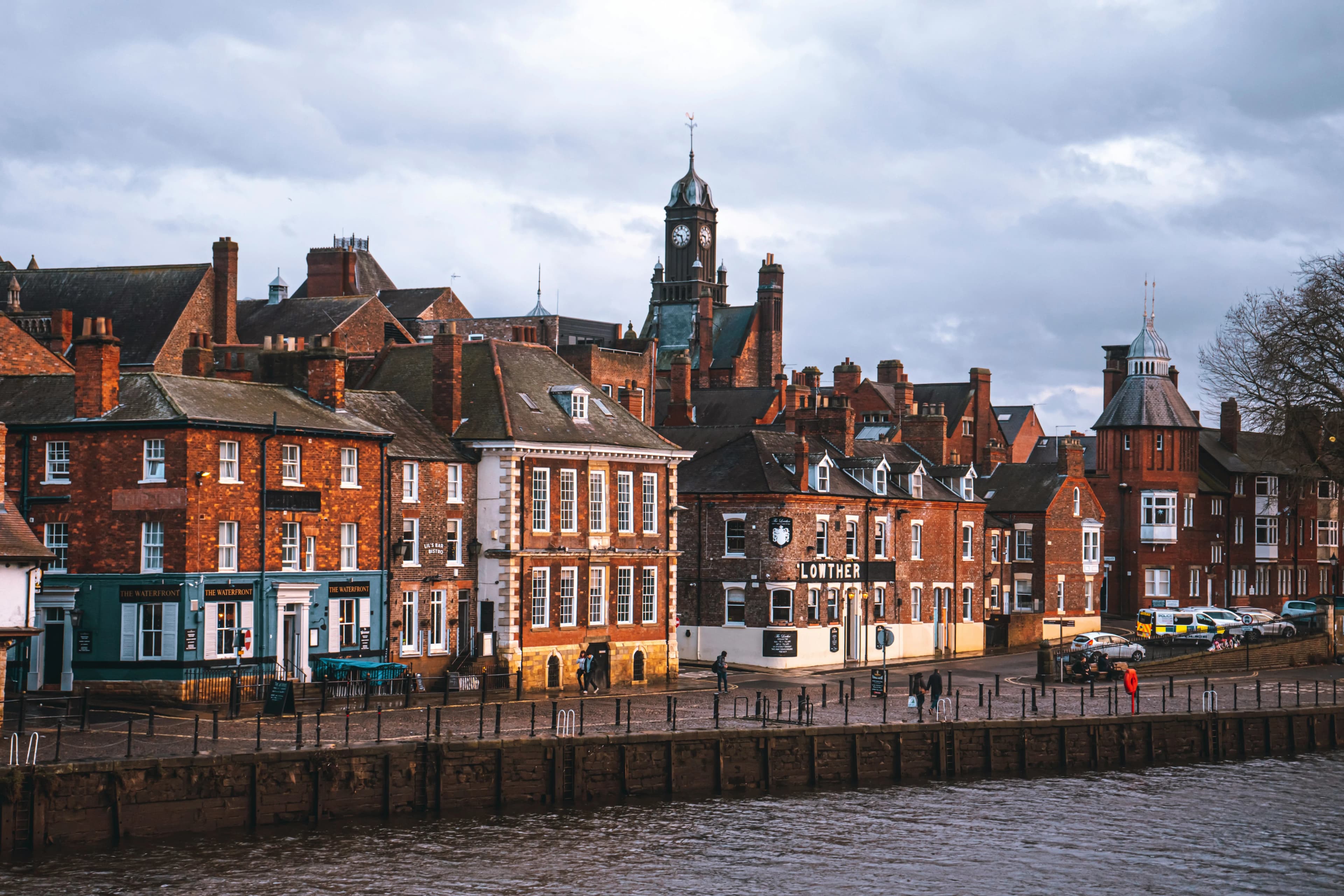 Historic brick buildings line the bank of the River Ouse, showcasing the city's charming architecture and a prominent clock tower.