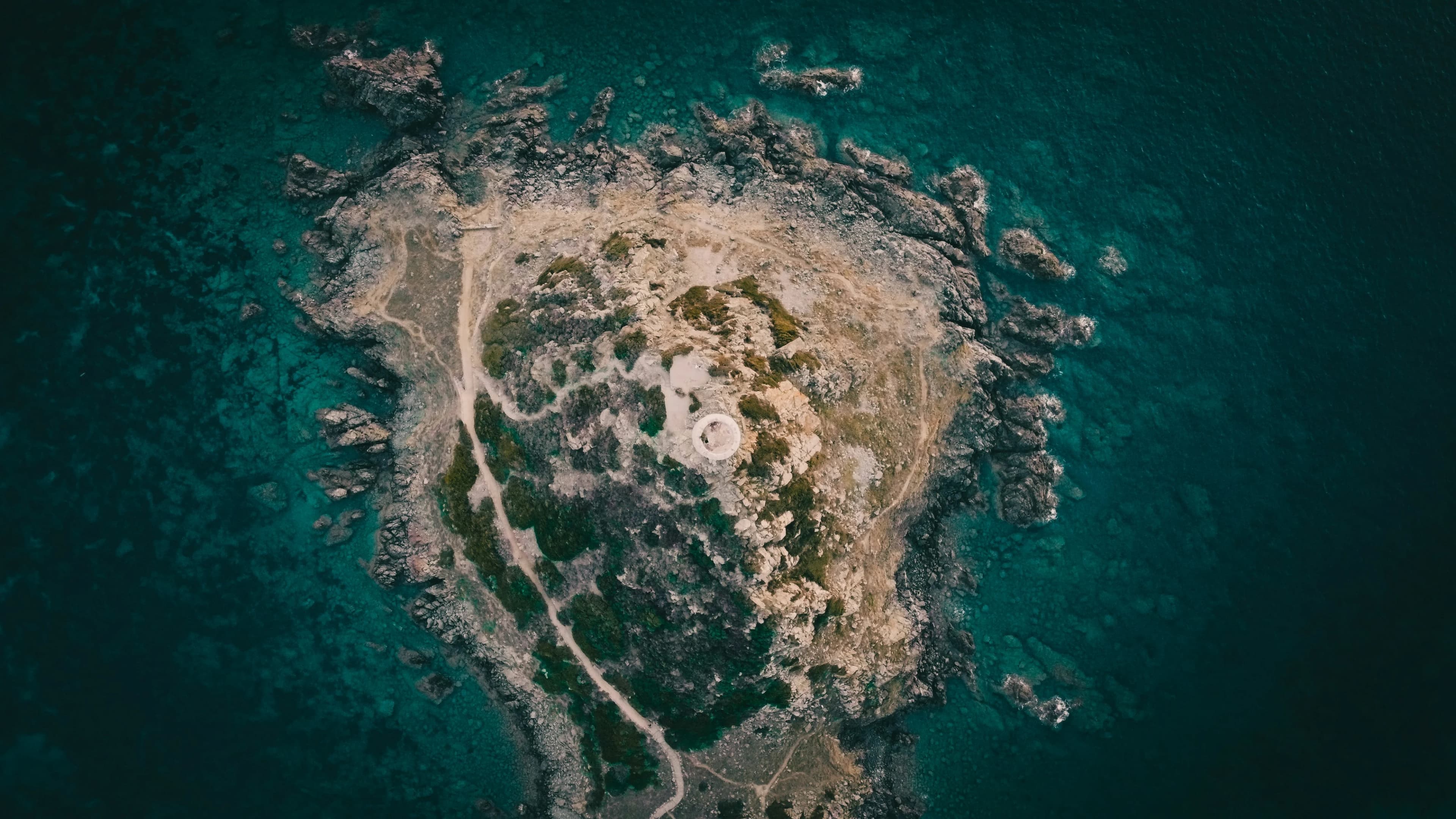 An aerial view captures a small, rocky island with a lighthouse, surrounded by the clear turquoise waters of the Mediterranean.