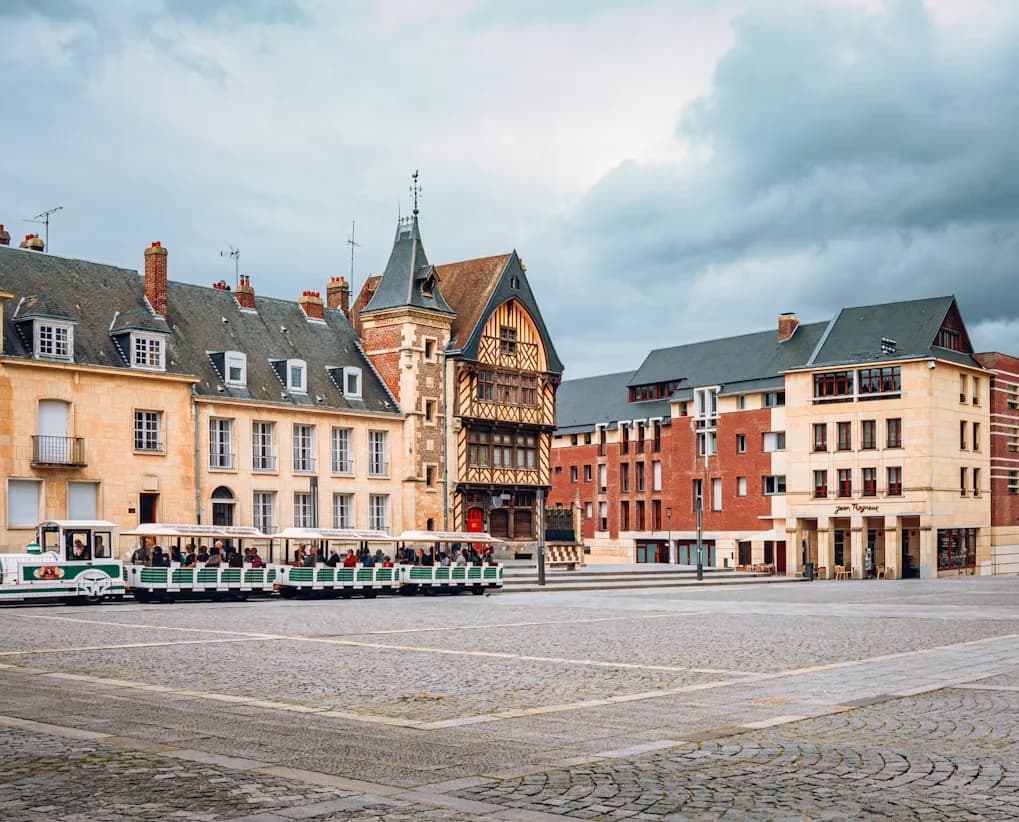 A small, white tourist train travels through a historic city square with traditional half-timbered buildings.