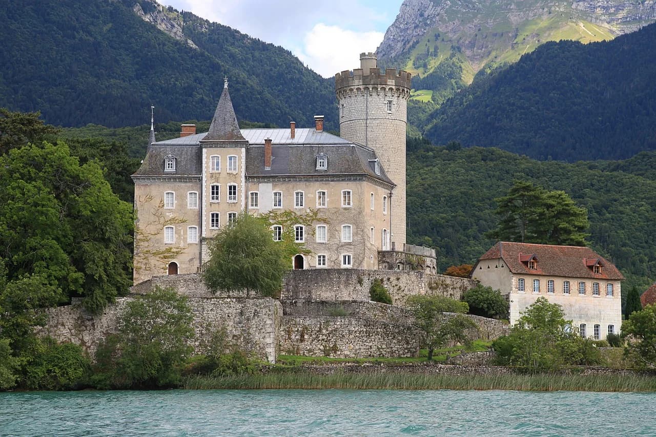The historic Château de Duingt, a fortress with a round tower, sits on the shore of Lake Annecy, surrounded by lush mountains.