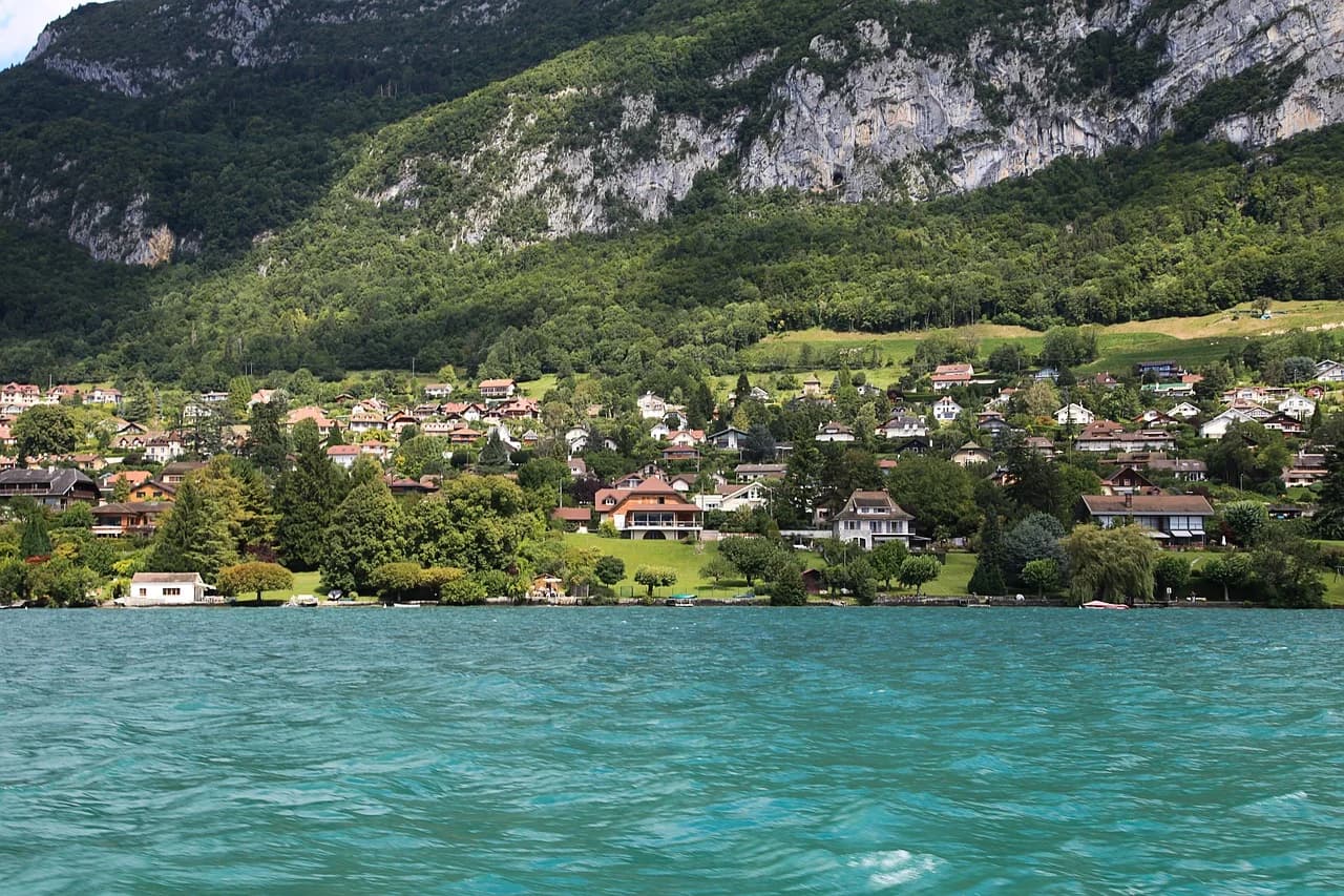 The beautiful turquoise waters of Lake Annecy are framed by a row of traditional houses and the lush green mountainside.
