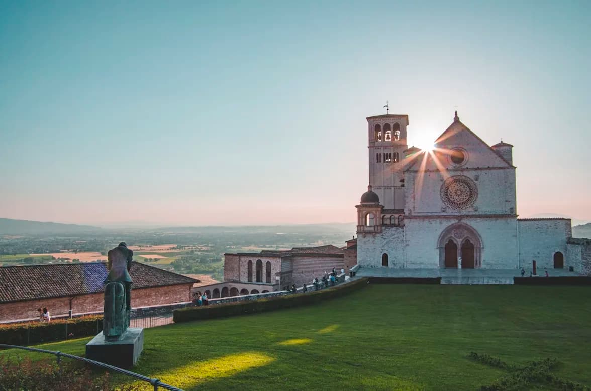 The Basilica of Saint Francis of Assisi stands on a hill overlooking the valley, with the golden sun setting behind it.