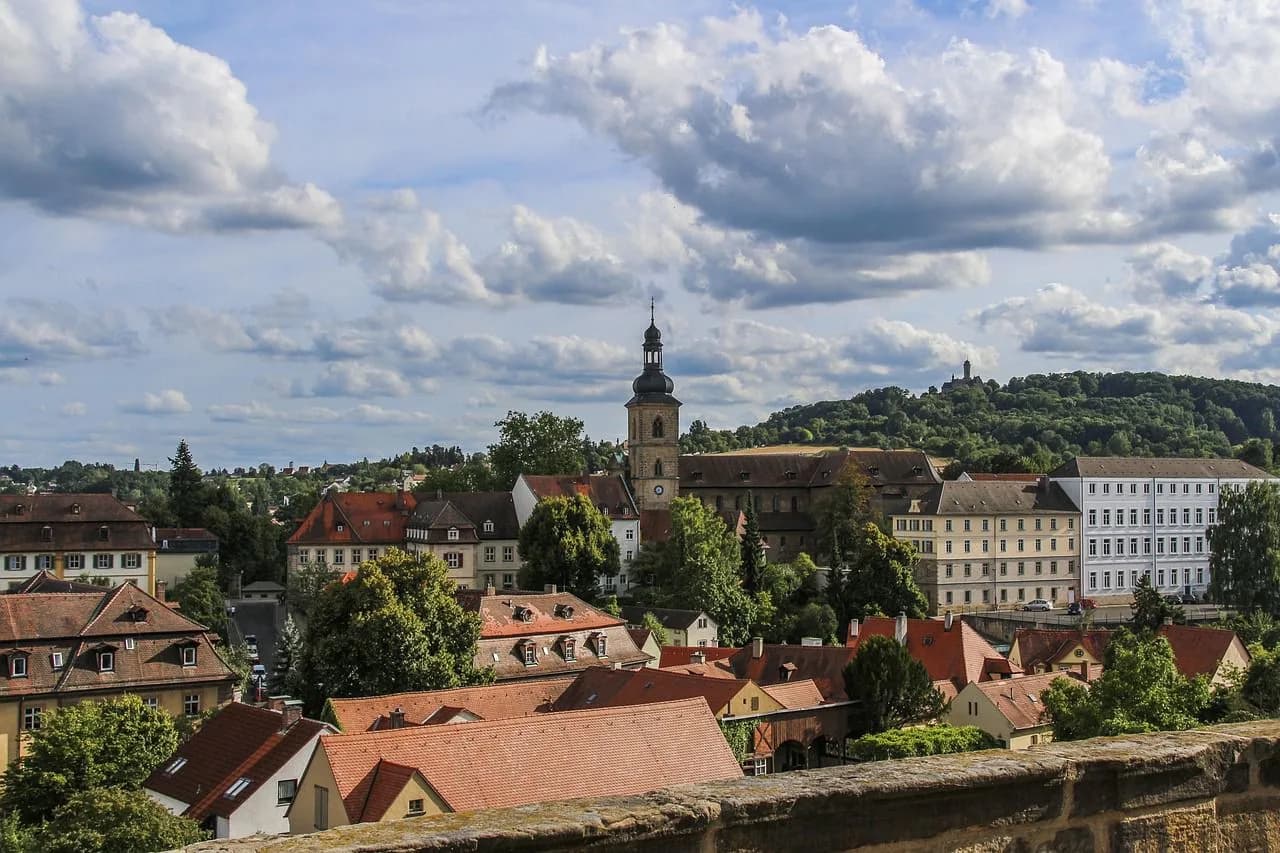 An elevated view captures the historic rooftops and church spires of Bamberg, with a green, forested hill in the distance.