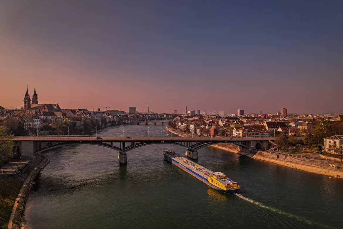 A cargo ship travels down the Rhine River, with a historic bridge and the city of Basel stretching along the banks.