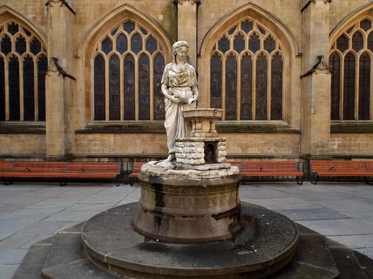 A classic Roman statue of a woman with a water pot sits on a fountain in front of the ornate windows of Bath Abbey.