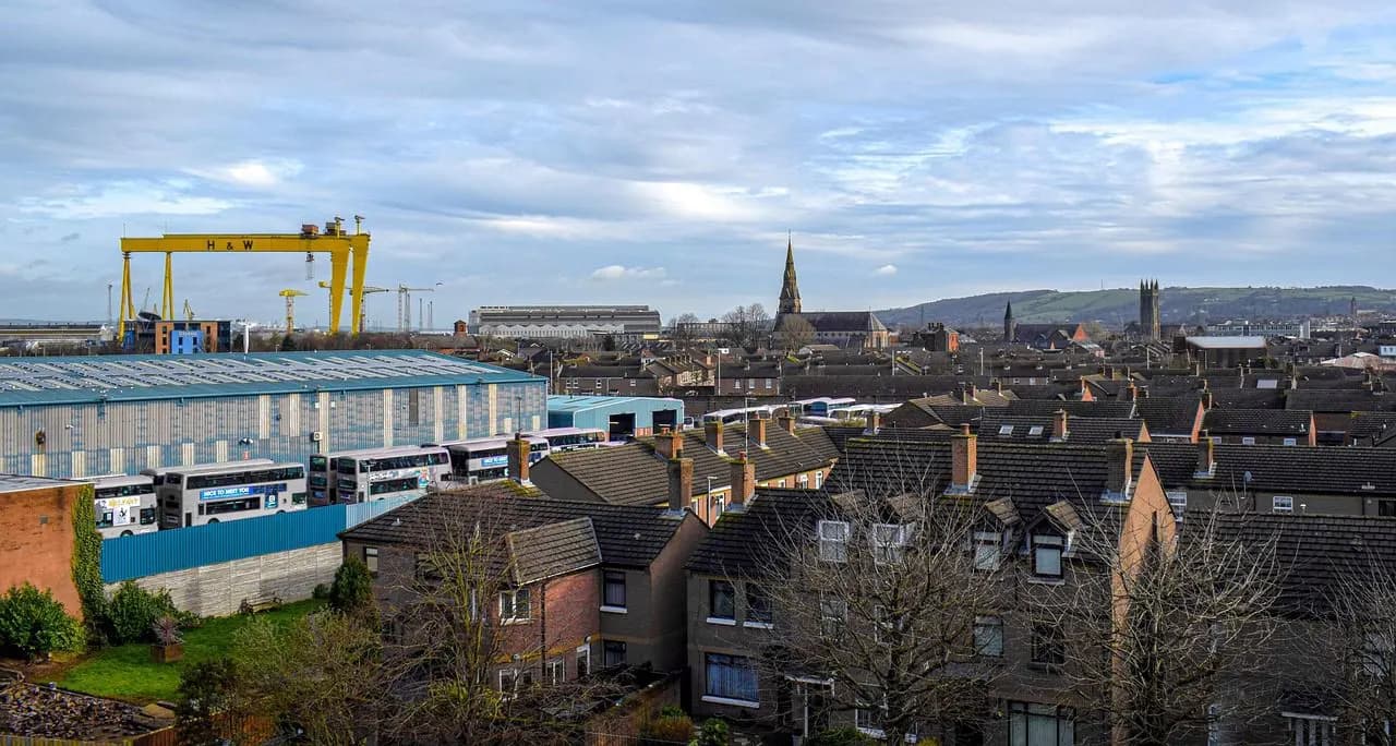 The iconic Harland and Wolff shipyard cranes stand in the distance, a symbol of Belfast's industrial heritage, overlooking residential rooftops and a church spire.