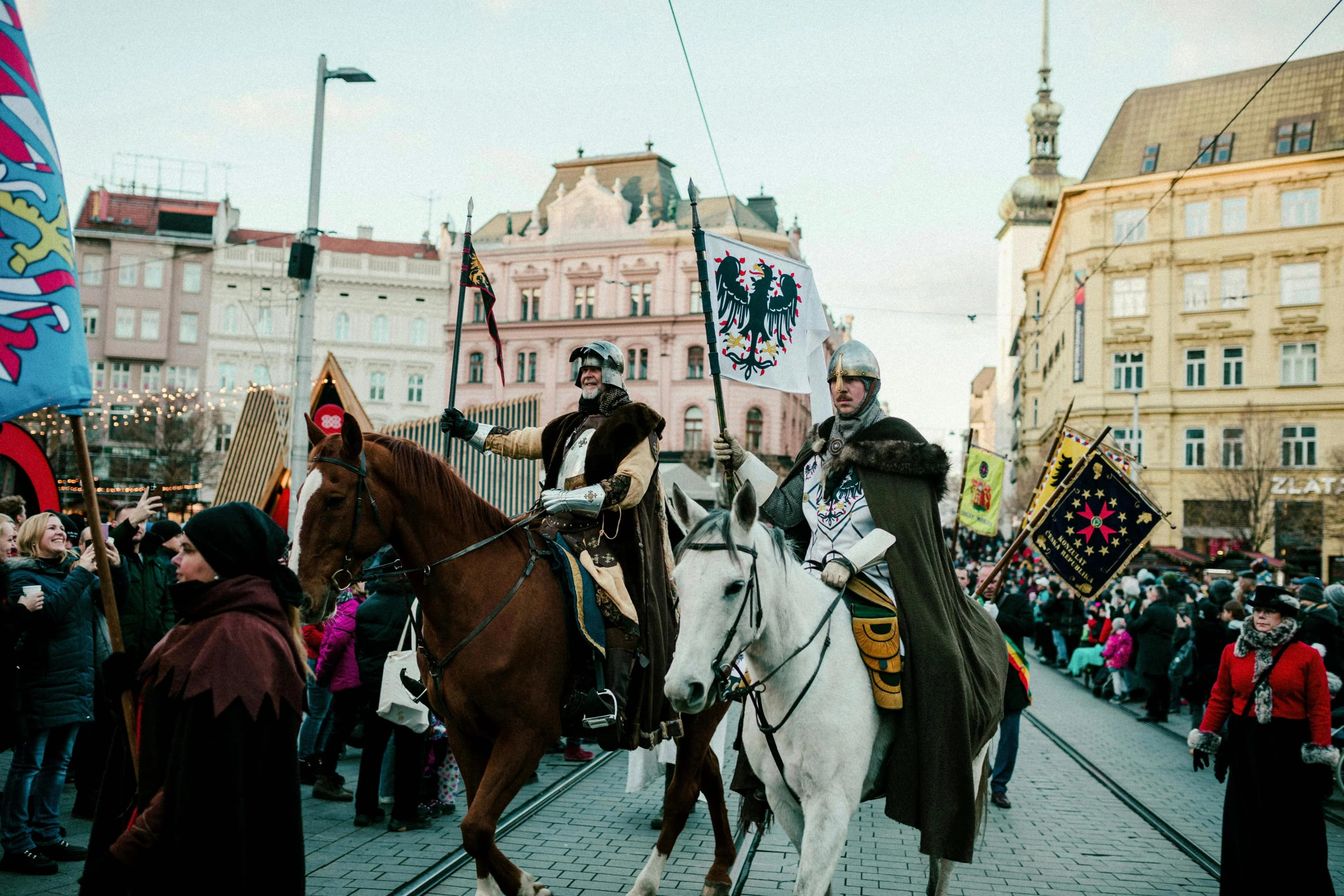 People dressed in historic costumes ride horses and carry flags in a city square, celebrating a traditional festival.