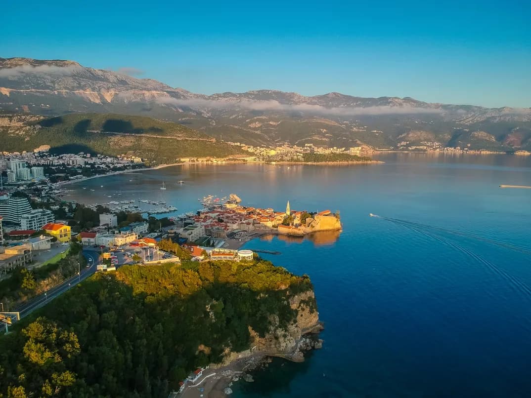 An aerial view of Budva shows the historic Old Town peninsula and the modern city, with the calm blue waters of the bay and dramatic mountains in the background.