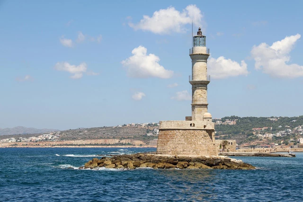 The iconic Venetian lighthouse of Chania stands on a stone breakwater, with the calm sea and the town in the distance.