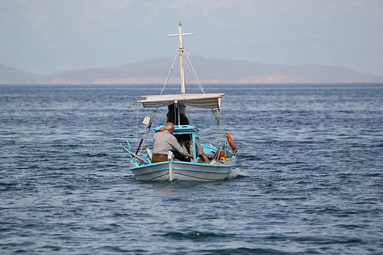 A small fishing boat with an elderly man at the helm sails on the calm, blue waters of the Aegean Sea.