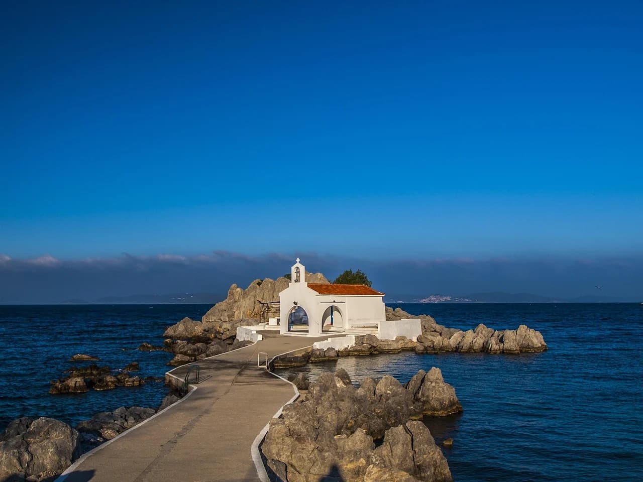 A small, white Greek Orthodox church is situated on a rocky outcrop, connected to the mainland by a winding walkway over the sea.