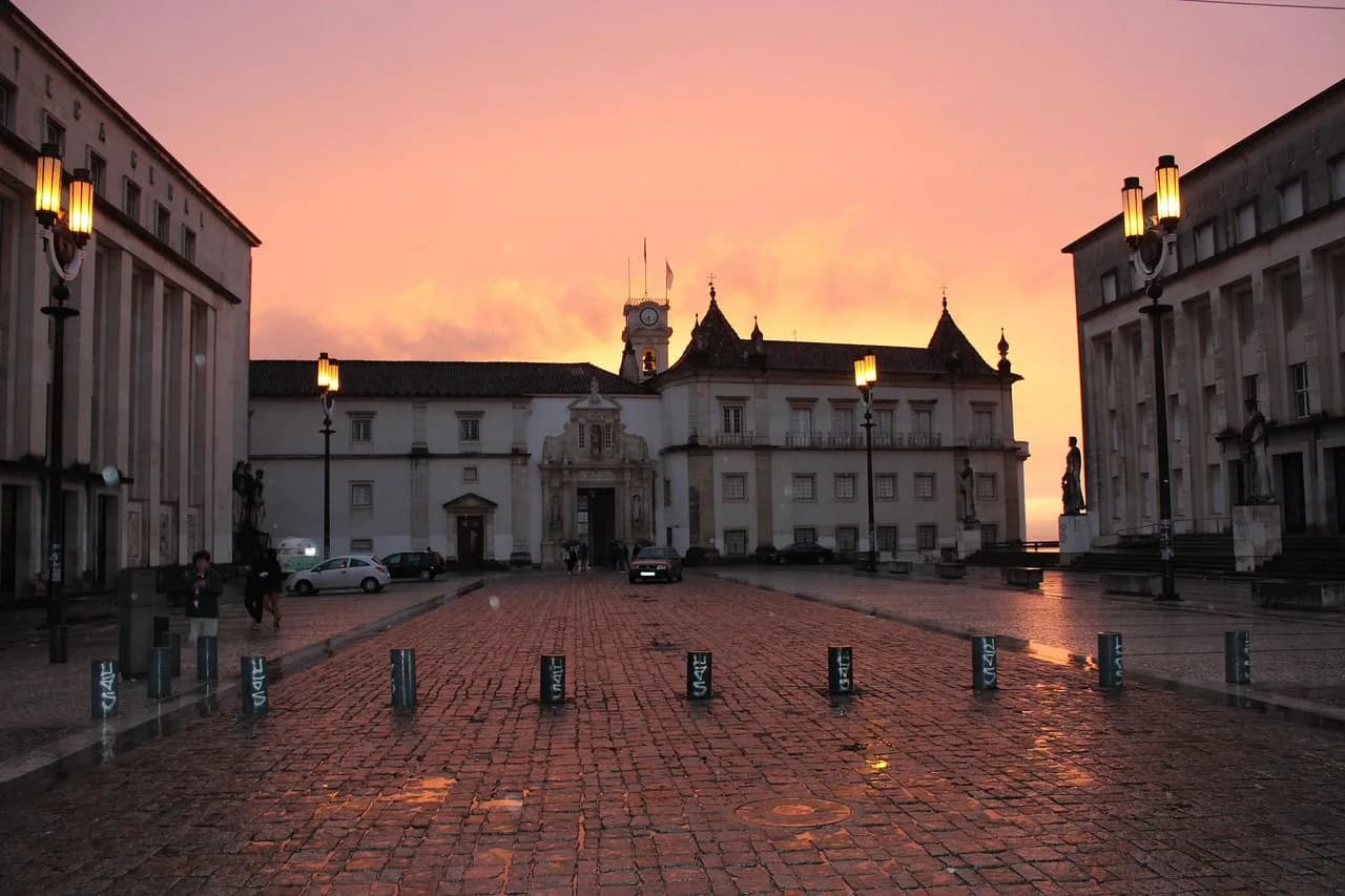 The grand central square of the University of Coimbra is bathed in the light of a sunset, with historic buildings and cobblestones creating a dramatic scene.