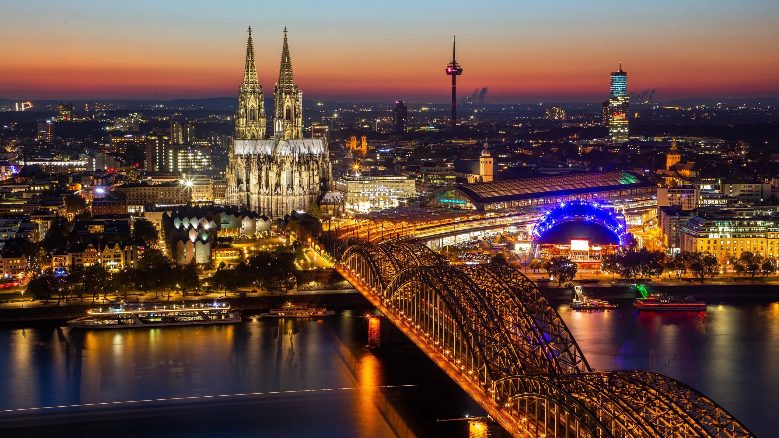 The illuminated Hohenzollern Bridge and Cologne Cathedral are beautifully reflected on the Rhine River at sunset, with the city lights glowing.