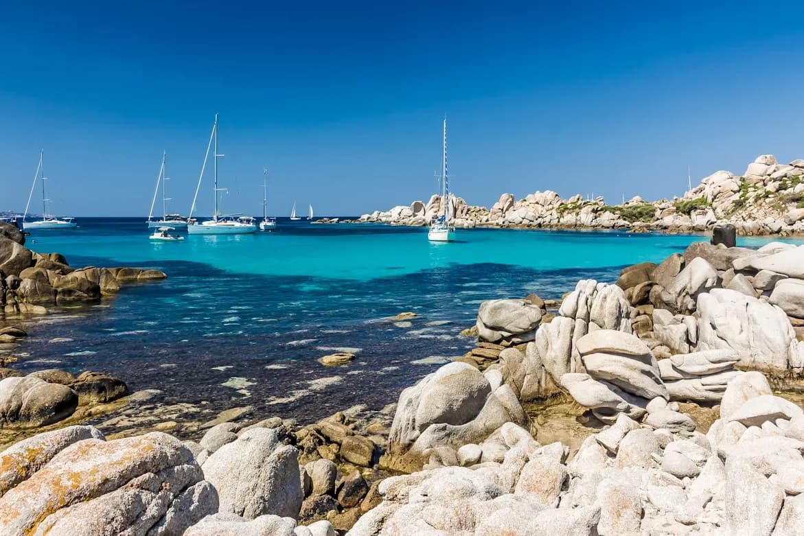 Sailboats are moored in a bay with shallow, turquoise water, surrounded by large, smooth, white rocks.