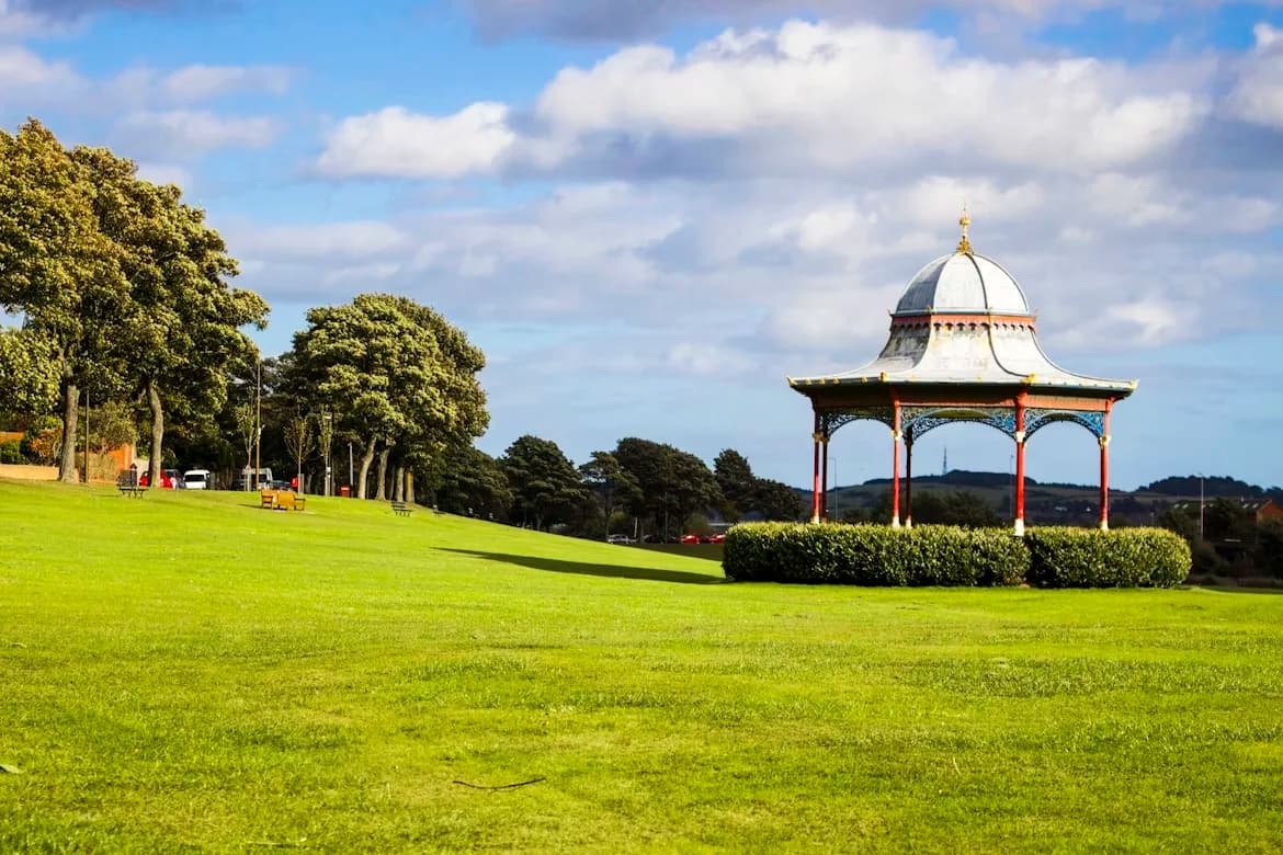 A historic bandstand sits on a large, green lawn, offering a serene view of the surrounding park.