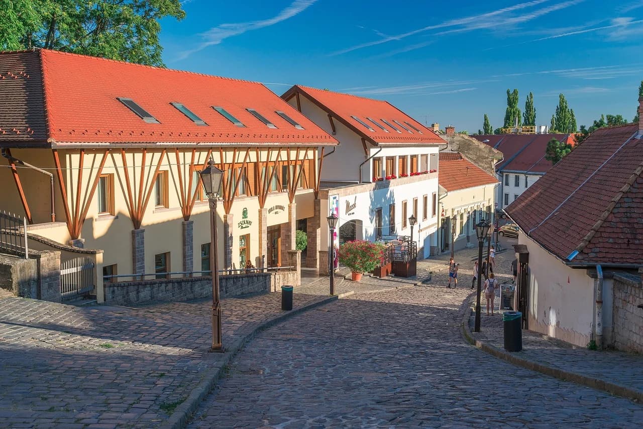 A quiet cobblestone street in Eger is lined with traditional buildings with red roofs, leading to a small square.