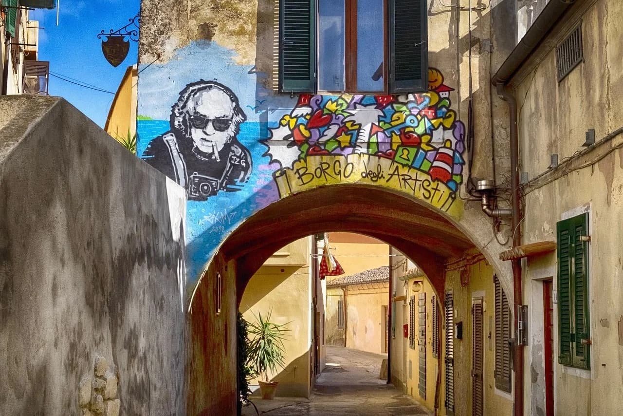 A colorful mural of an artist with a camera and a graffiti tag is painted on a building, creating a vibrant scene in a narrow Italian alleyway.