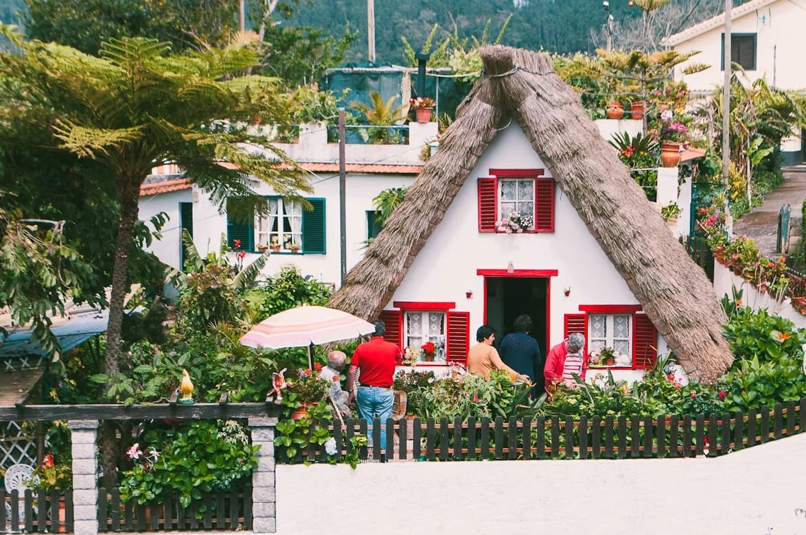 A unique traditional cottage with a thatched roof and red-framed windows is nestled in a beautiful garden setting.