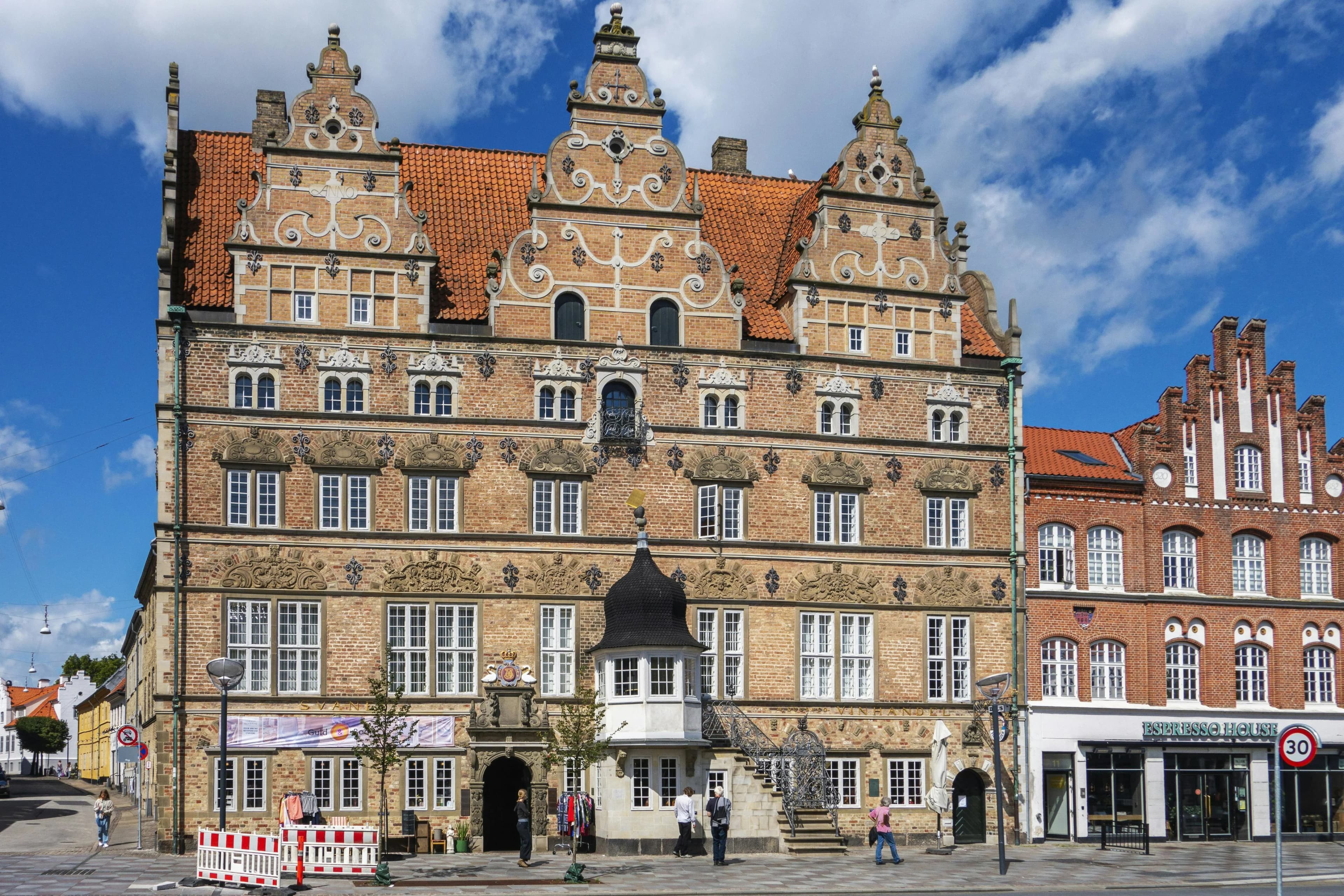 A historic, gabled building with ornate decorations stands in a city square under a bright blue sky.