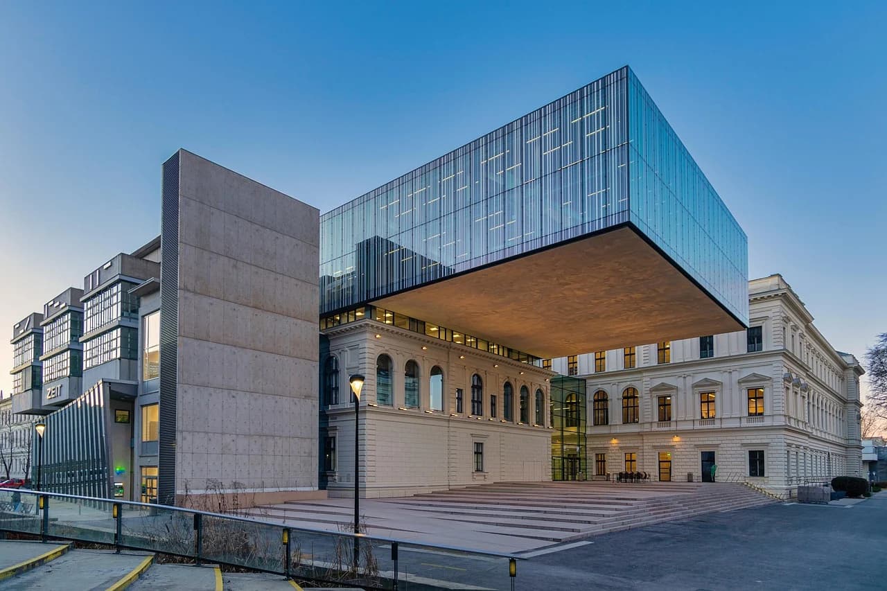 A striking modern glass structure with a cantilevered roof is seamlessly integrated with a historic building, showcasing a blend of old and new architecture.