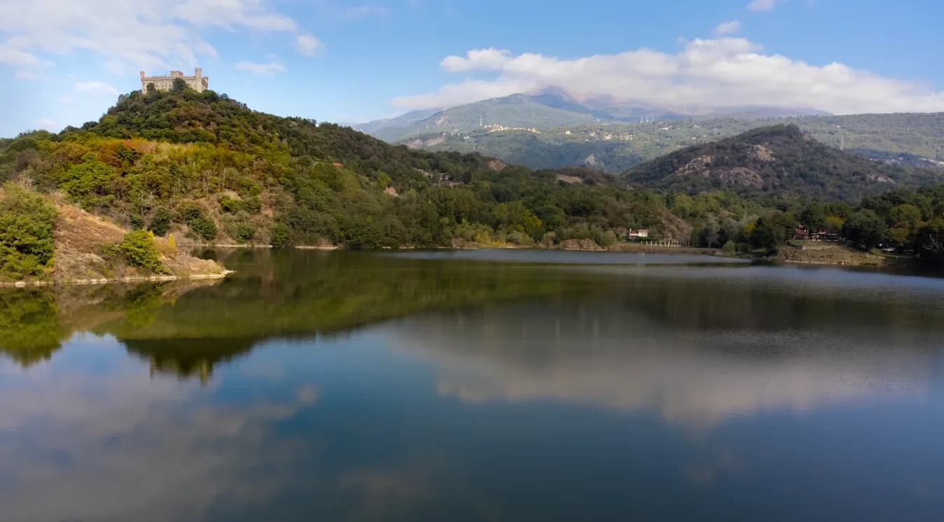 A medieval castle sits atop a lush green hill, overlooking a serene lake that perfectly reflects the surrounding landscape and mountains.