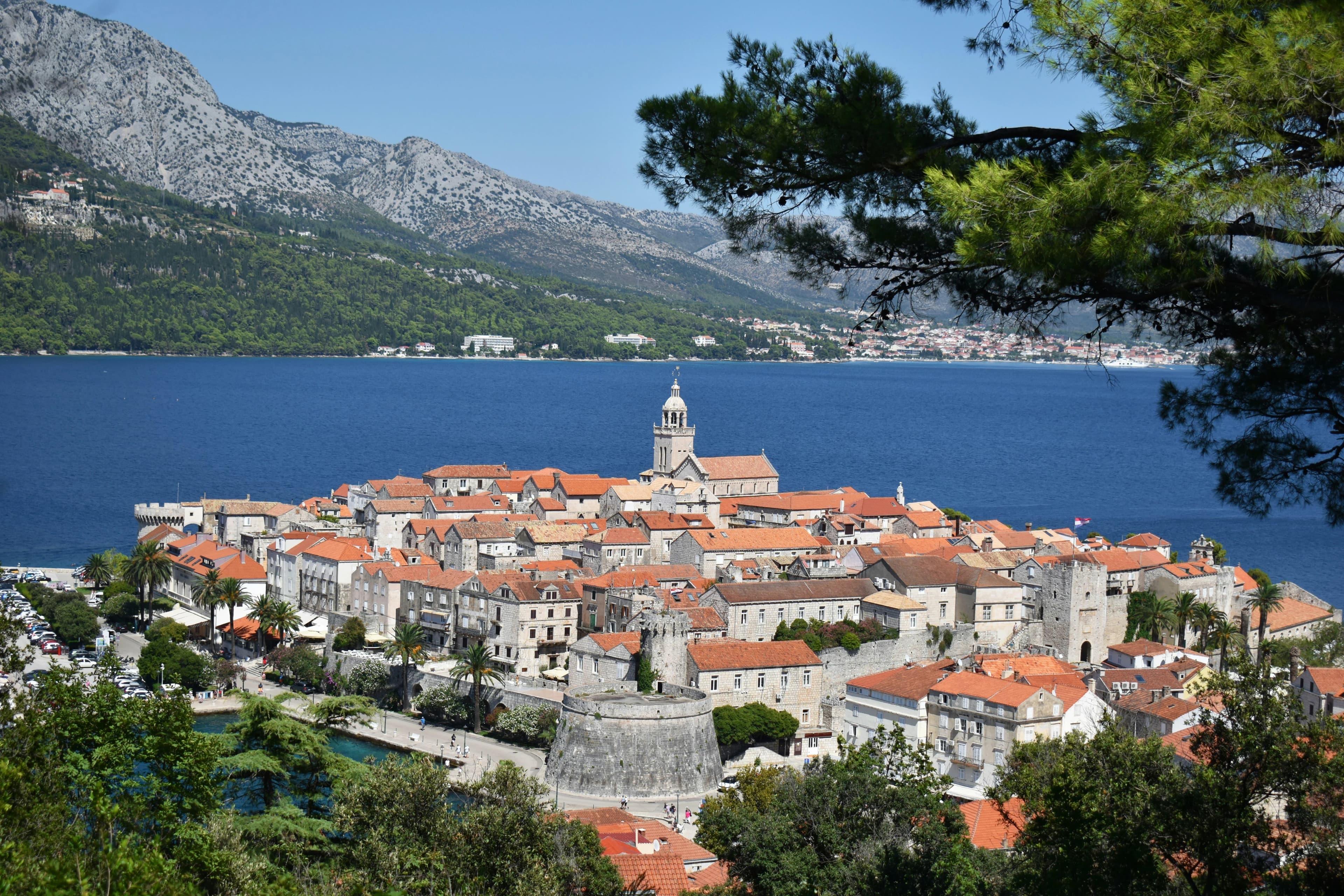 A large sailing ship with a red and white sail is a beautiful sight in a bay with a historic town and mountains in the background.