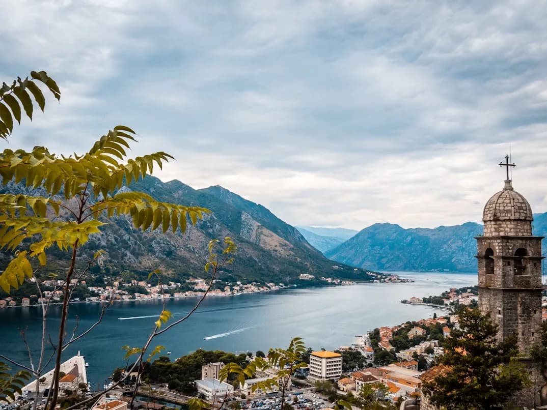 A historic stone bell tower sits in the foreground, providing a frame for the majestic Bay of Kotor and the surrounding mountains.