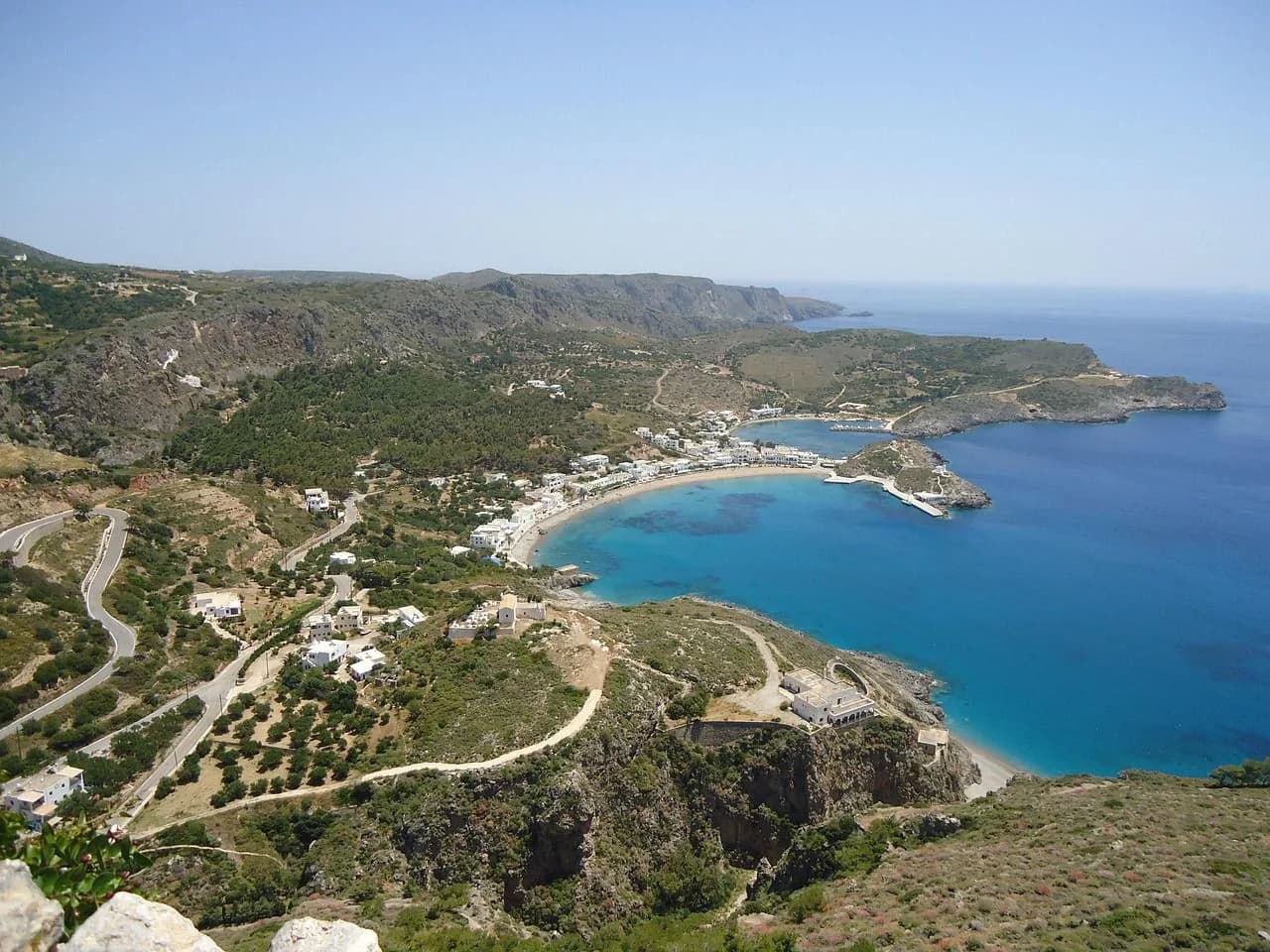 A winding road leads down to a beautiful, crescent-shaped bay, with white buildings and a clear blue sea.