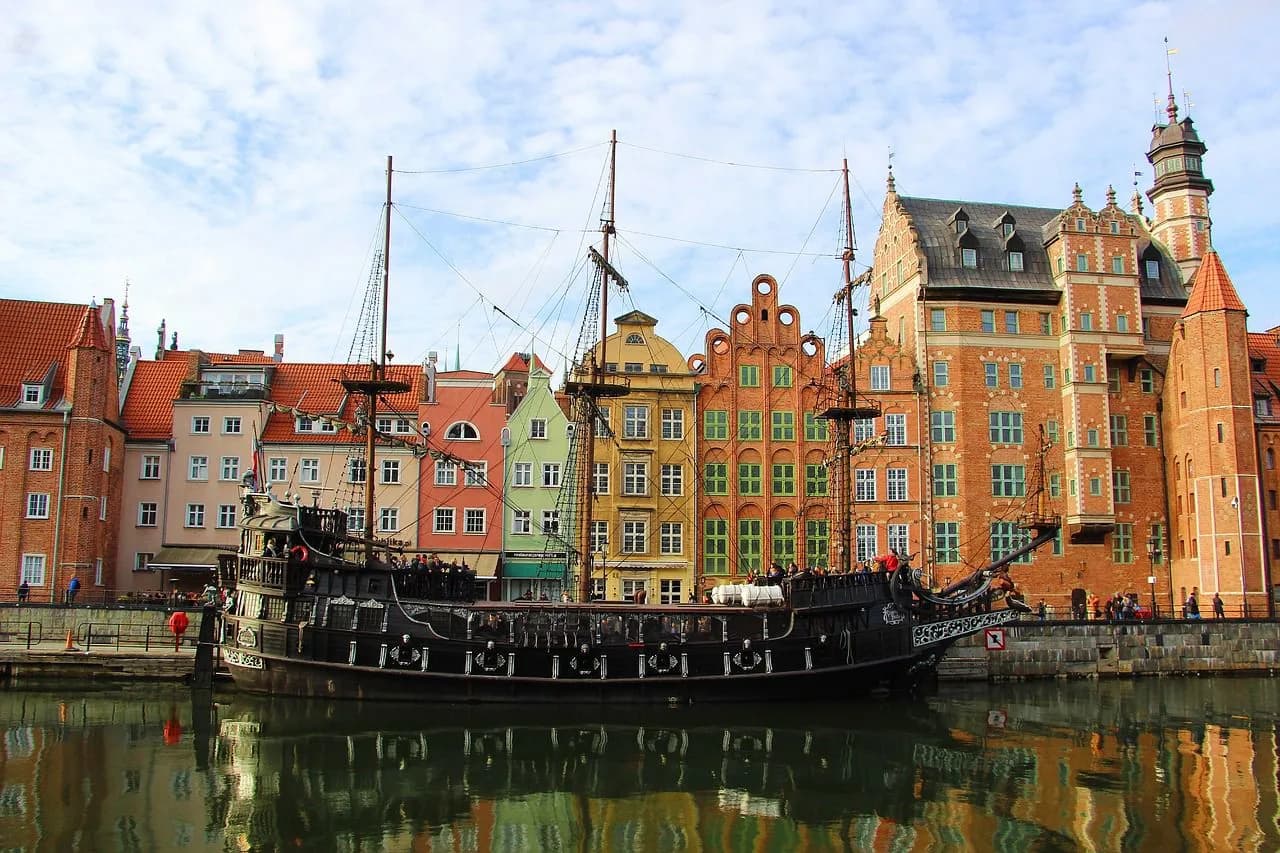 A large, historic sailing ship is moored at the pier in Gdańsk, with the colorful facades of the city's historic buildings in the background.