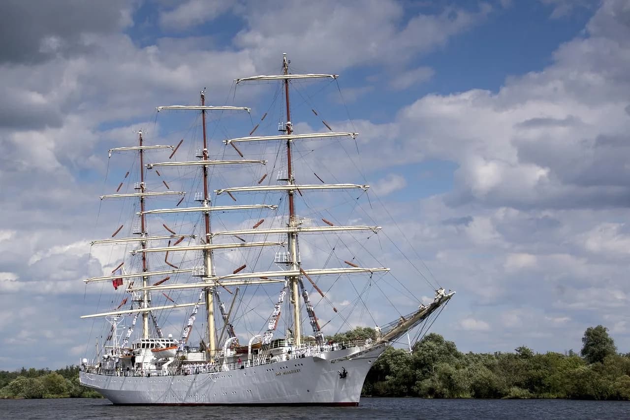 A large, white sailing ship with intricate rigging sails on a lake, surrounded by a green shoreline.
