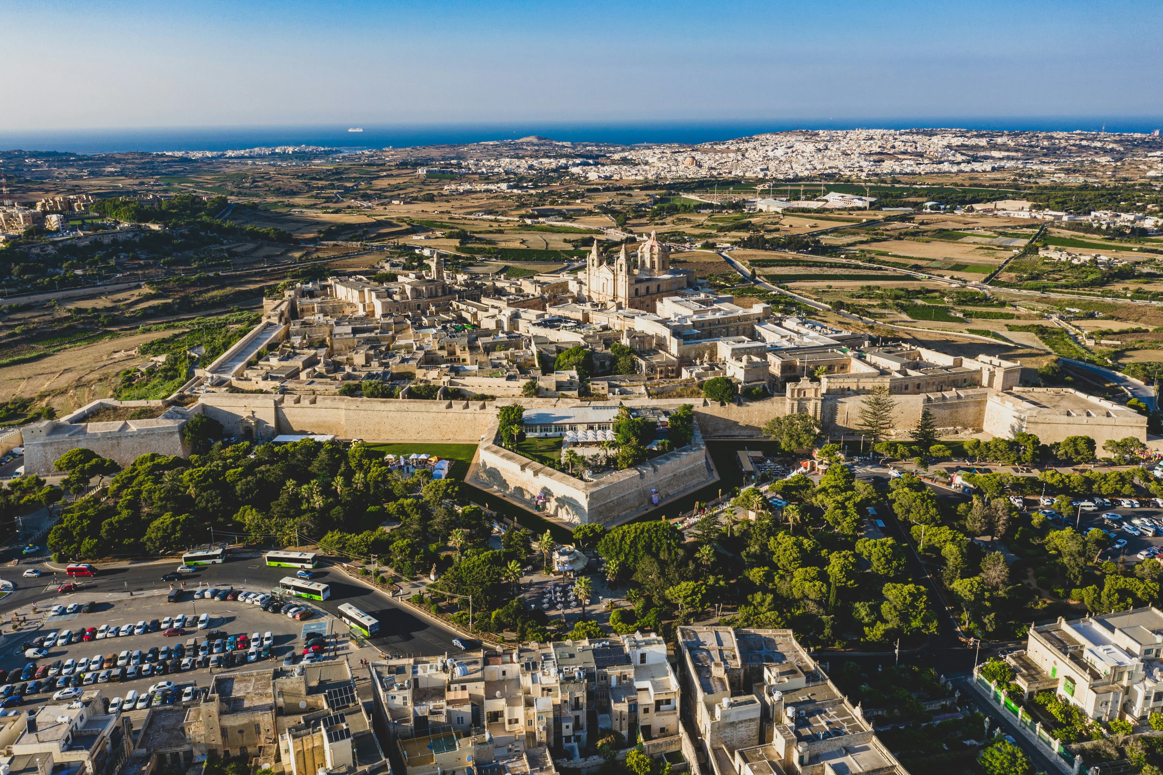 An aerial view captures the walled city of Mdina, with its intricate buildings, and the surrounding green landscape stretching to the sea.