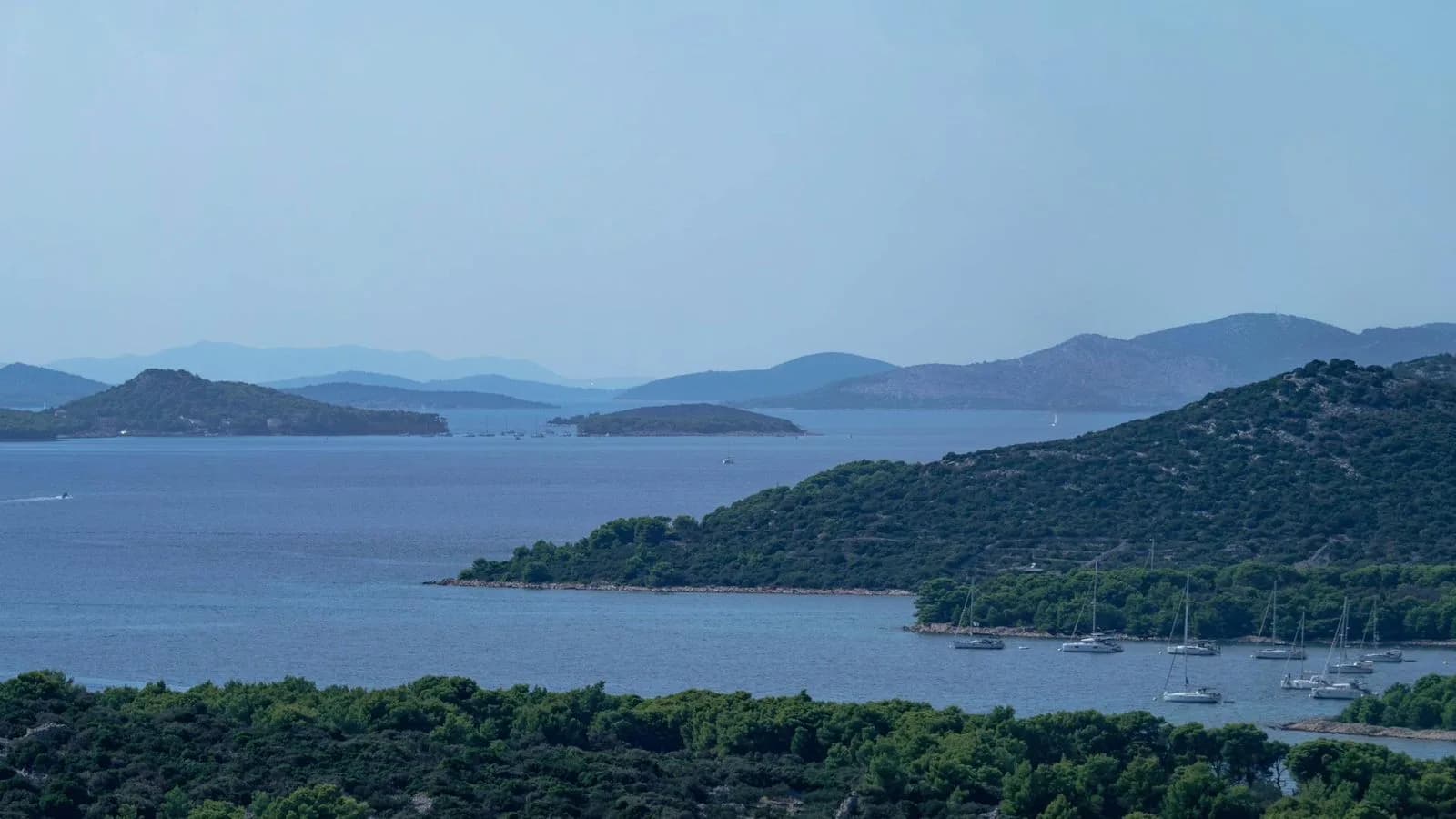A serene bay is filled with sailboats and yachts, with a series of islands and green hills in the distance.