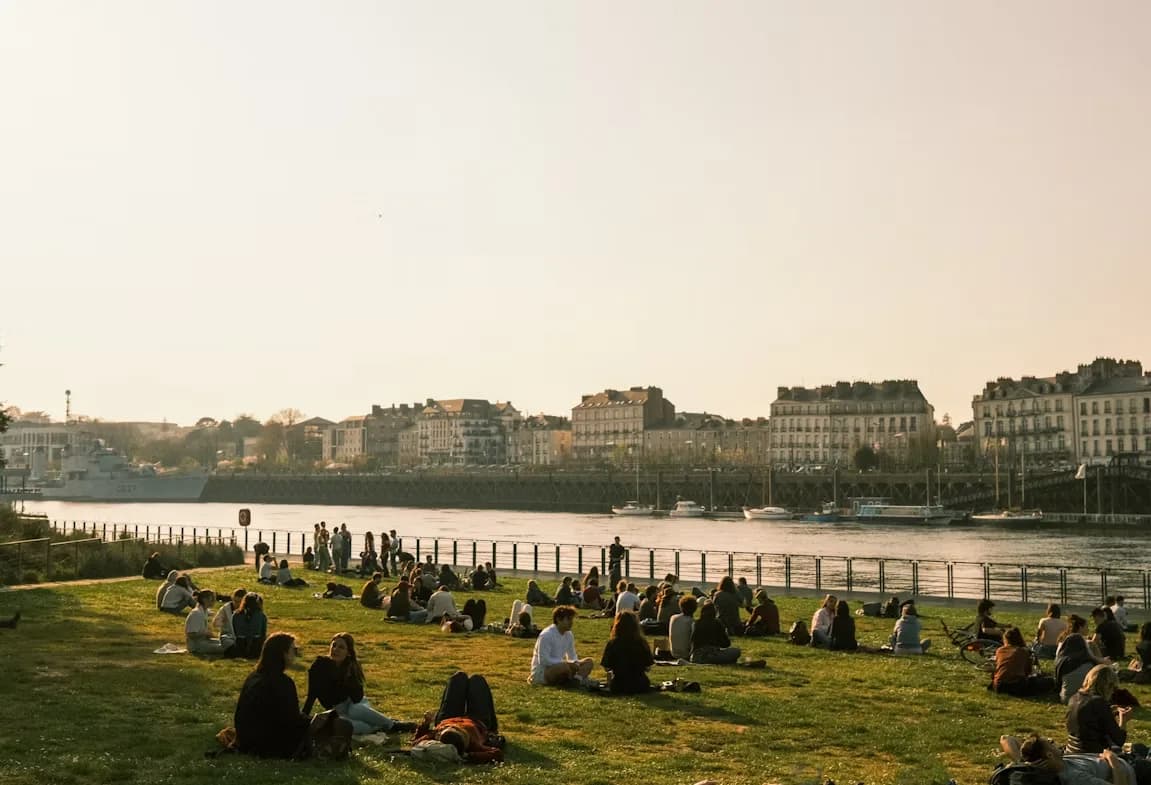 People relax on a grassy bank by a river, with the historic buildings and a military ship in the background.