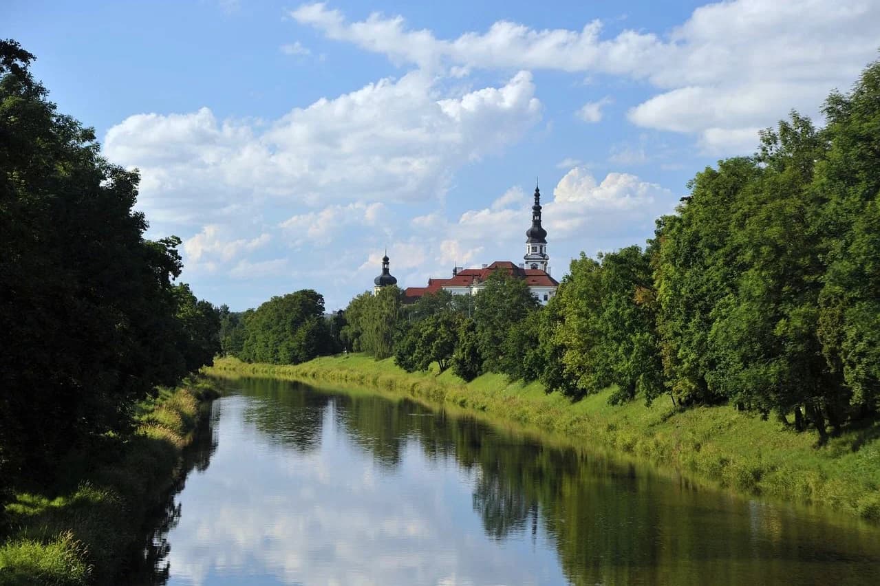 A tranquil river flows through a lush green landscape, with a historic church and a monastery in the background.