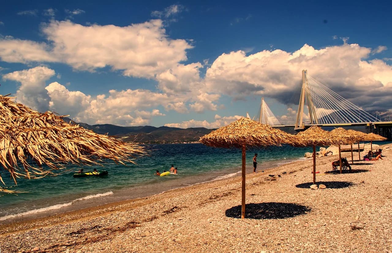 Thatched umbrellas and sun loungers line a pebble beach, with a view of the Rio-Antirrio Bridge in the distance.