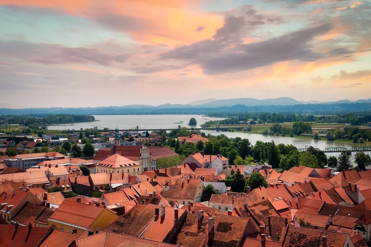 A panoramic view of the city of Ptuj shows a sea of red-tiled rooftops, with the Drava River and a bridge in the distance at sunset.