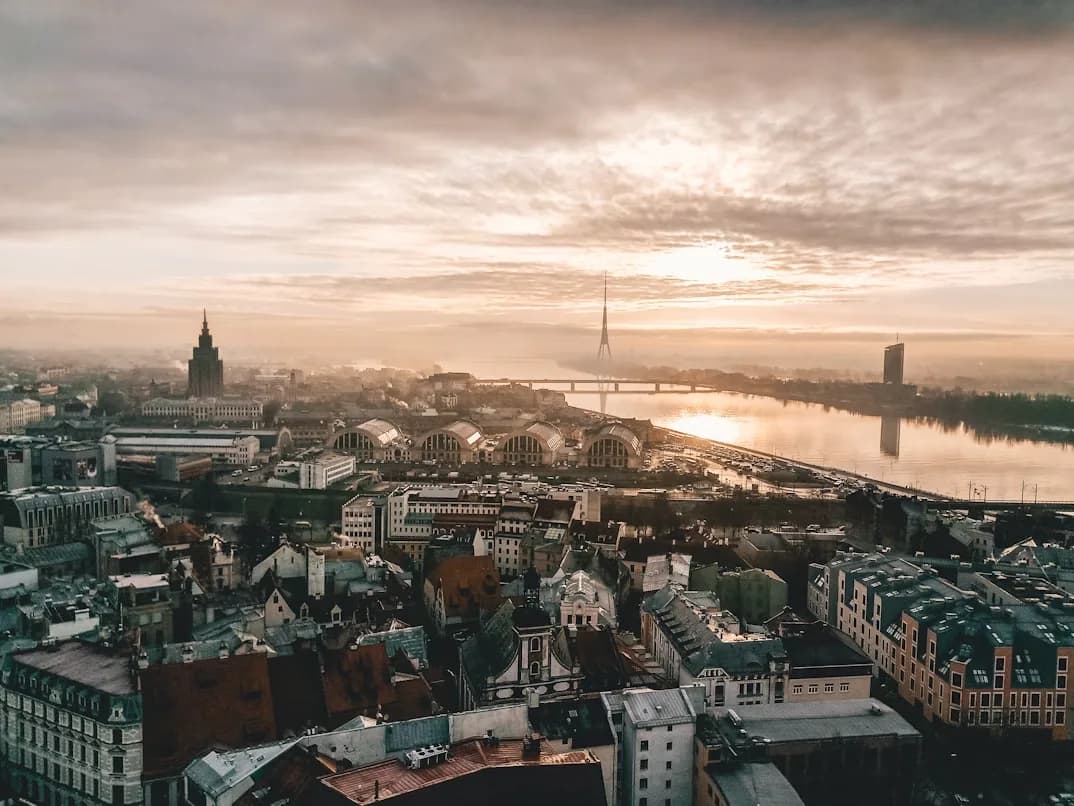 An aerial view of Riga at dawn shows the city's rooftops and a river with a bridge, with the sky in soft shades of orange and pink.