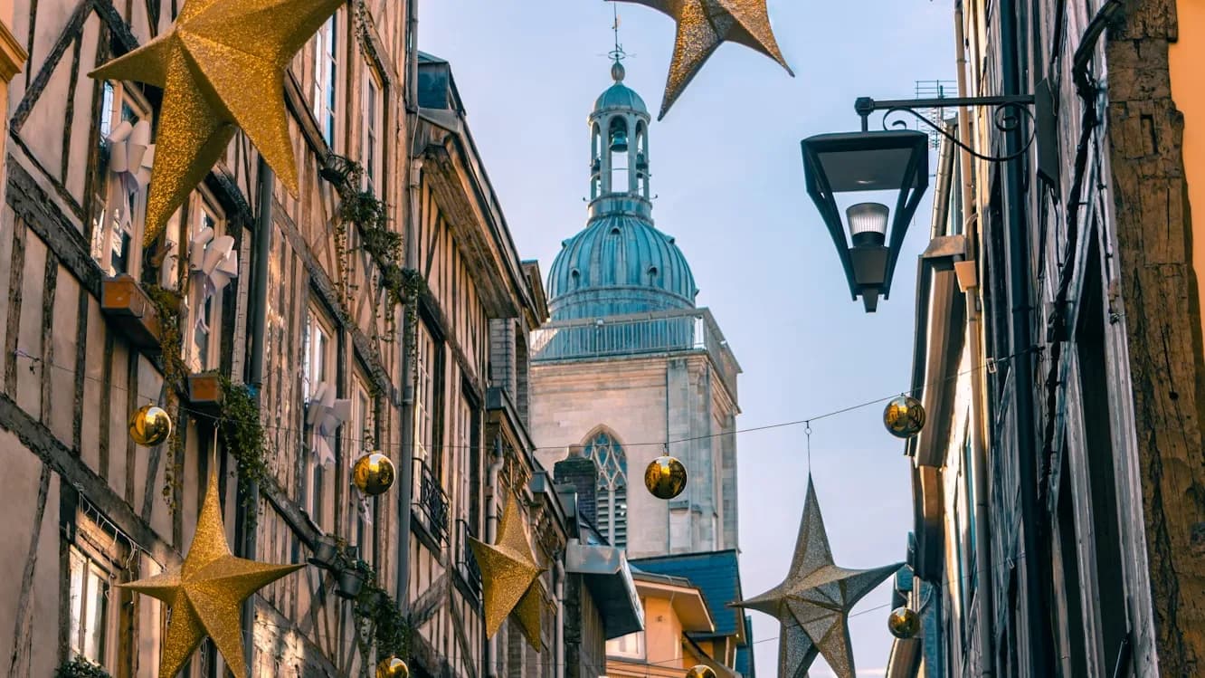 A historic street in Rouen is decorated with large golden star ornaments and festive garlands, with a church steeple in the background.