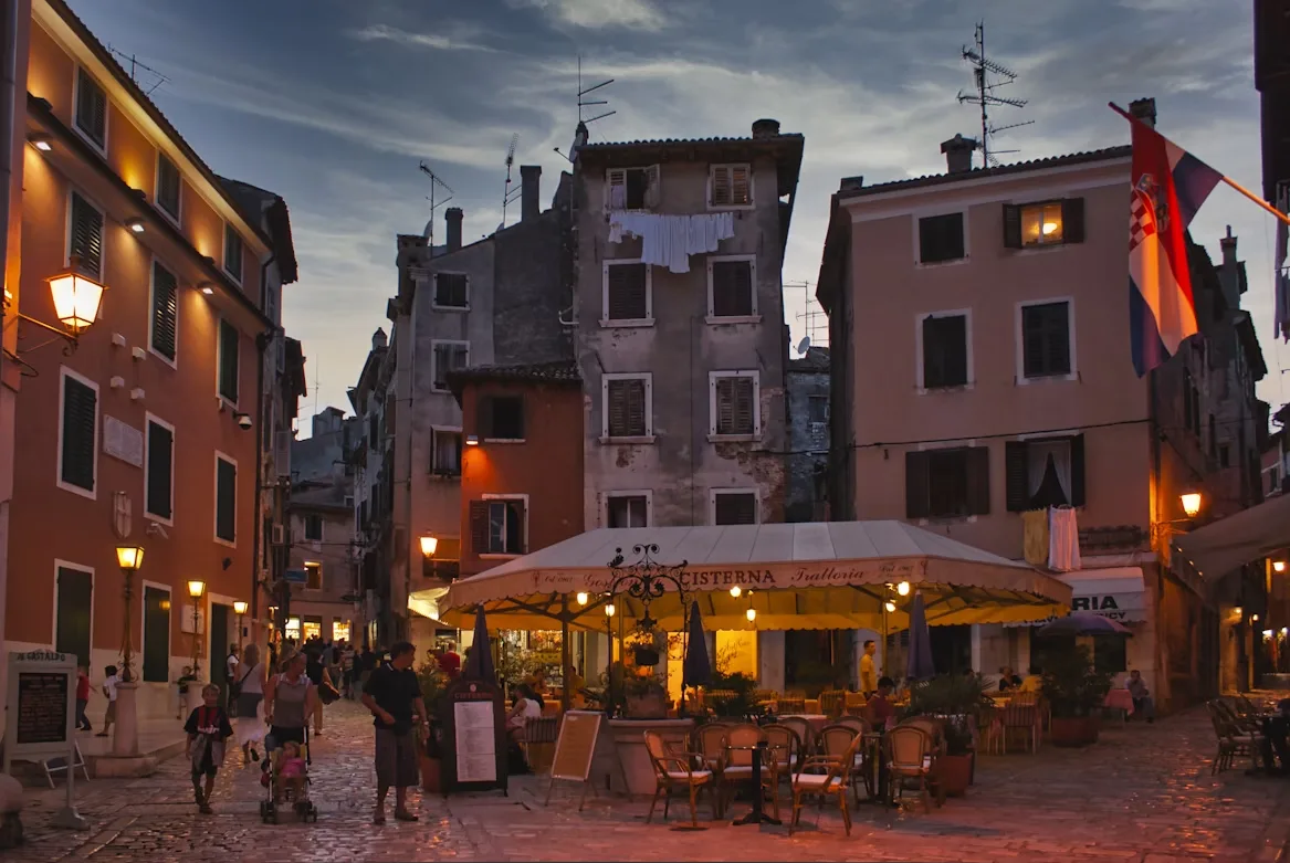 A narrow, cobblestone street in Rovinj is lined with historic stone buildings and bustling outdoor cafes.