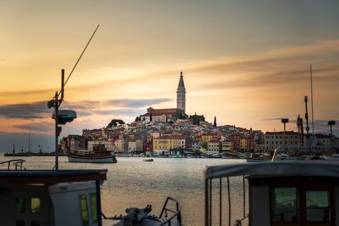 The skyline of Rovinj is beautifully illuminated at sunset, with the bell tower of St. Euphemia and boats in the harbor.