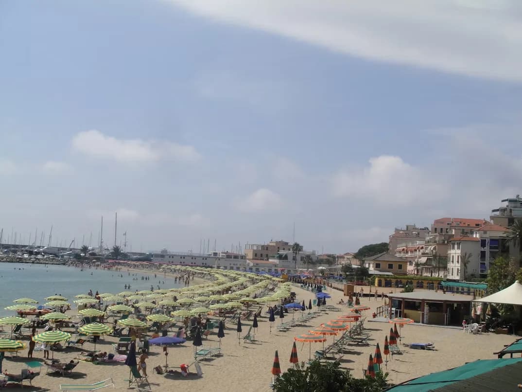 A wide, sandy beach is filled with rows of striped umbrellas and people relaxing, with the sea and a mountain in the background.