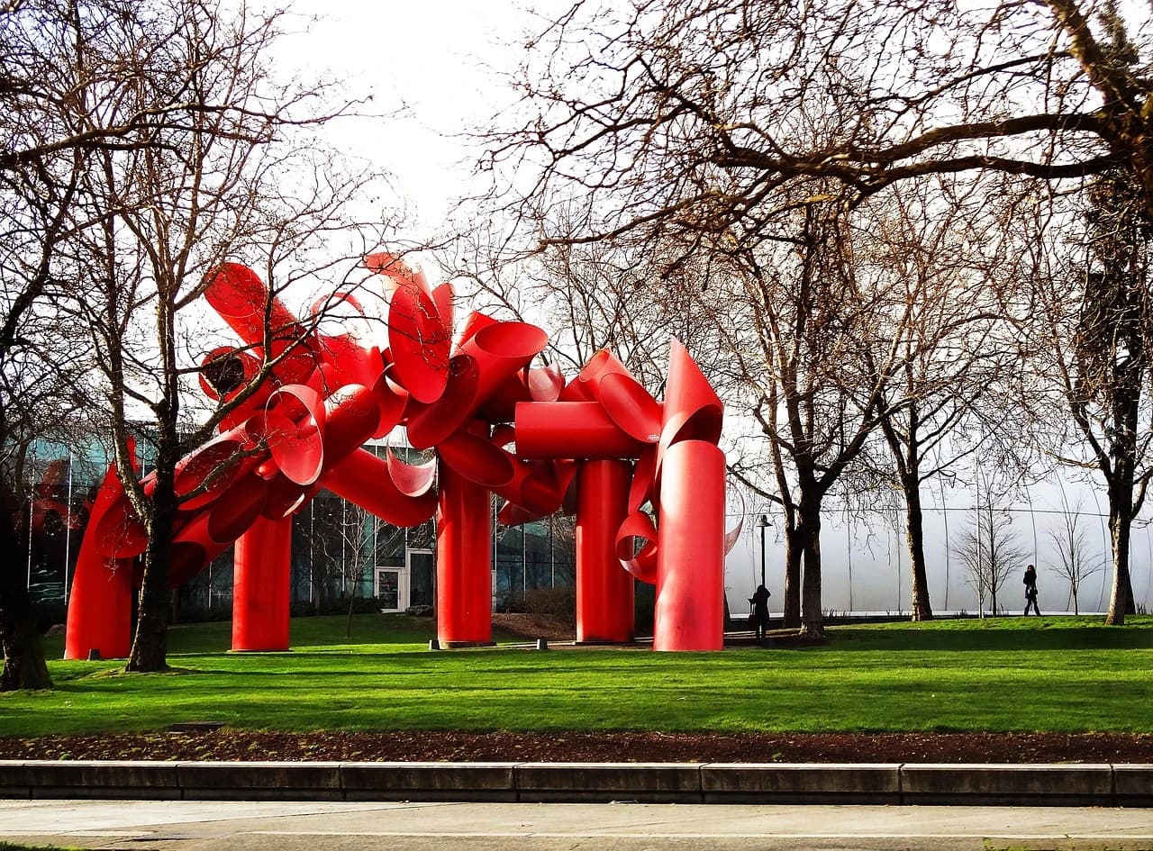 A large, abstract red sculpture stands in a grassy park, with a line of bare trees in the background.