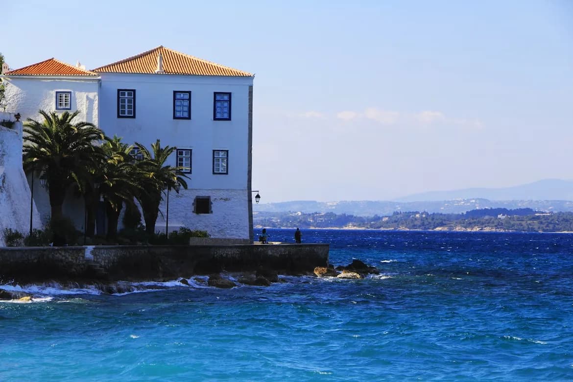 A traditional white house with a red-tiled roof stands on the rocky coastline, with palm trees and a view of the sea.