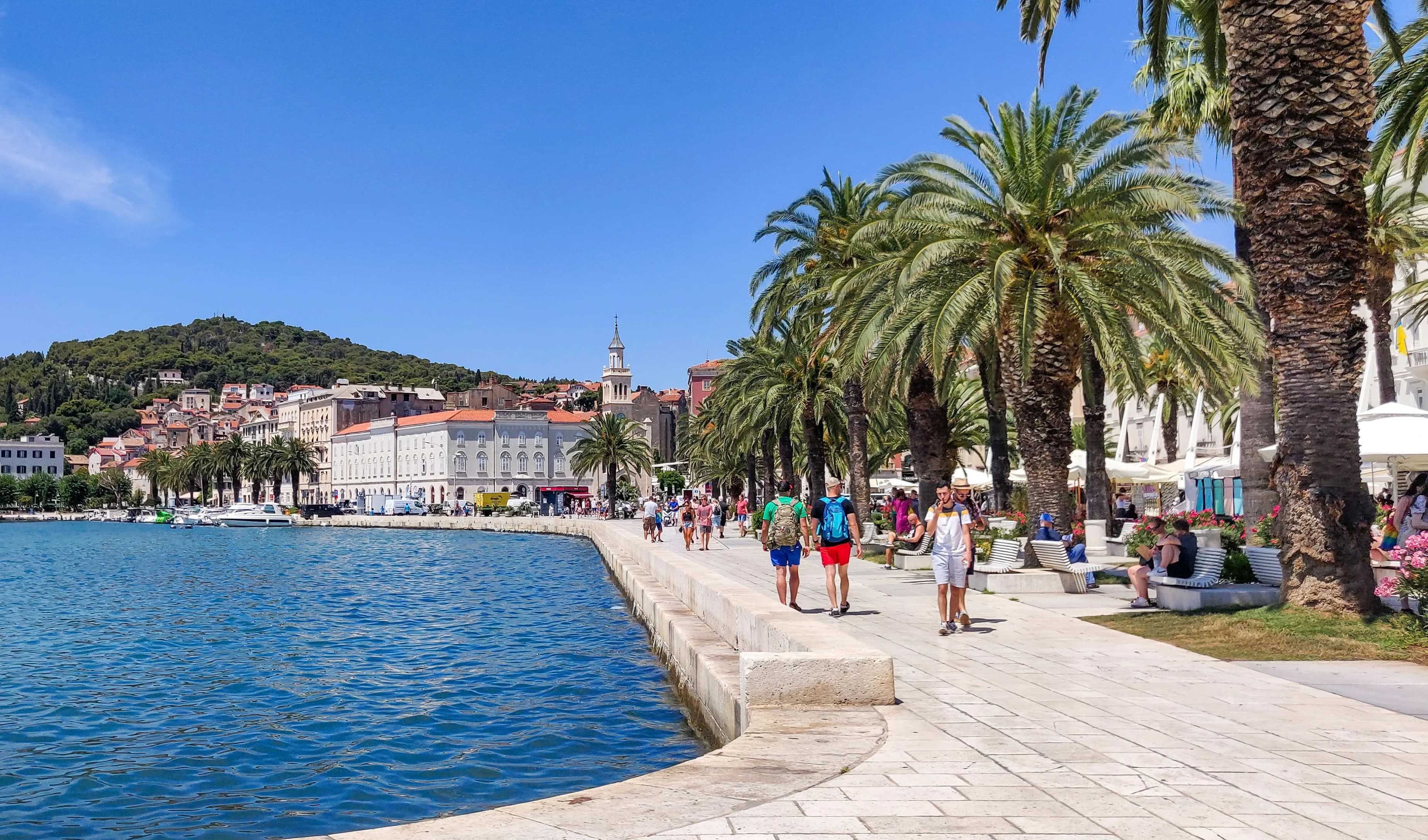 People strolling along the sun-drenched waterfront promenade in Split, lined with palm trees and outdoor cafes.