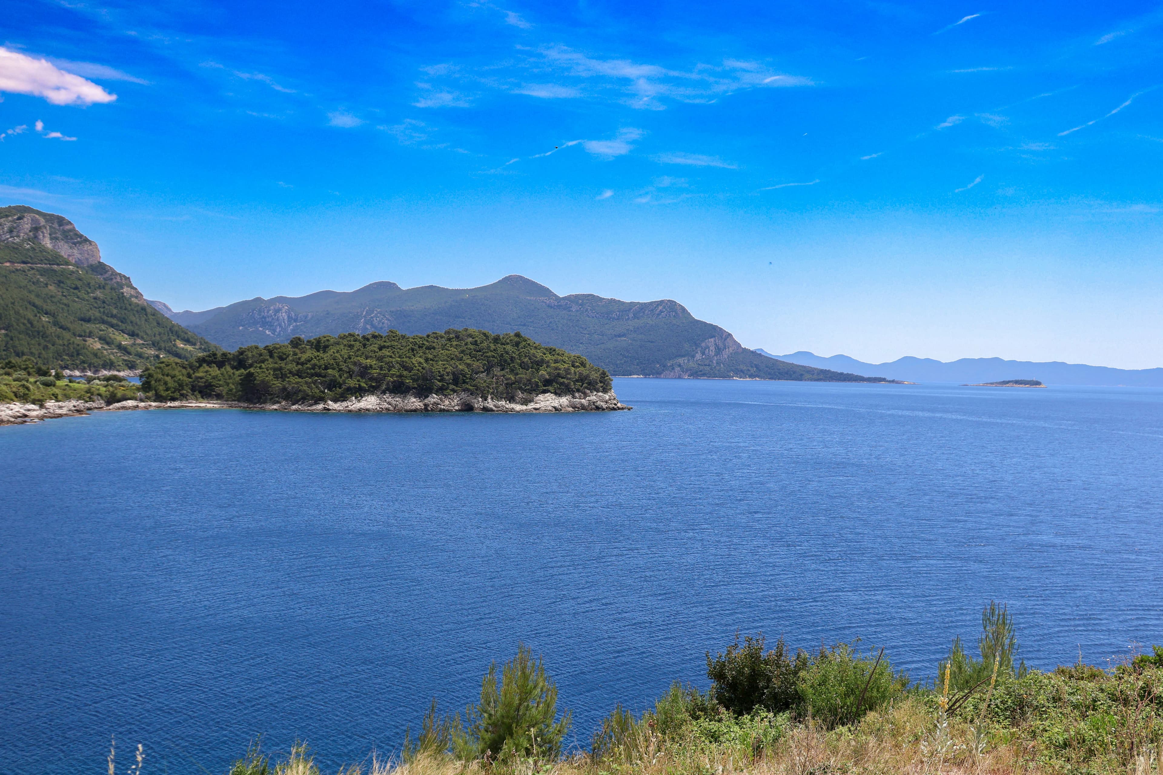 A scenic bay is surrounded by green hills and a few small islands, with the deep blue Adriatic Sea stretching to the horizon.
