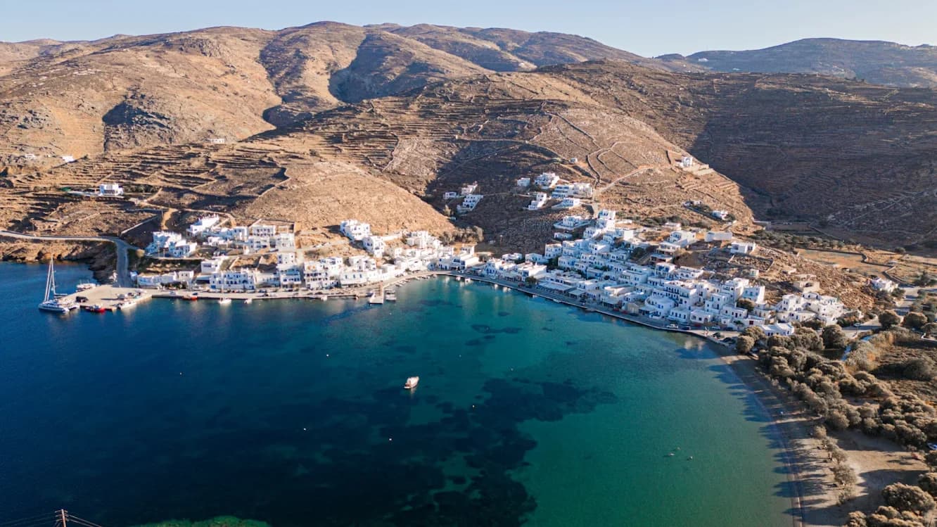 A quiet harbor in Tinos is filled with fishing boats and small yachts, with traditional white buildings and a historic bell tower in the background.