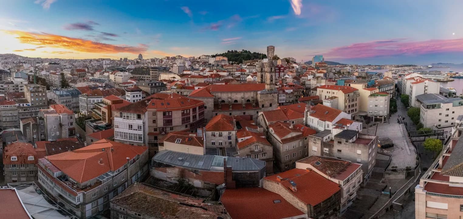 A panoramic view of the Vigo city skyline shows a dense cluster of red-tiled roofs, with the sea and distant hills in the background at sunset.