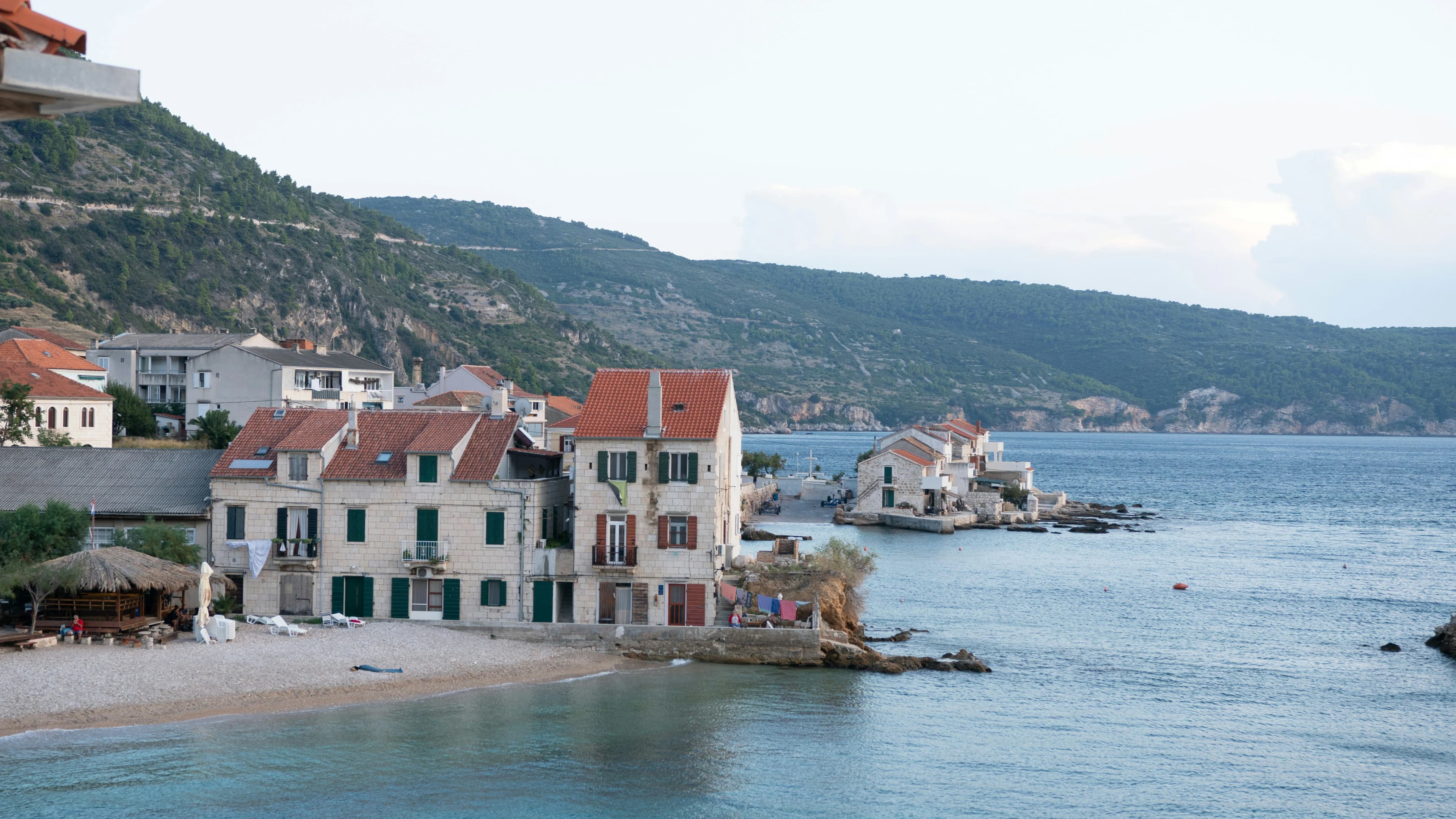 The tranquil bay is lined with a small, sandy beach and traditional buildings with red-tiled roofs, with a hillside and a mountain in the background.