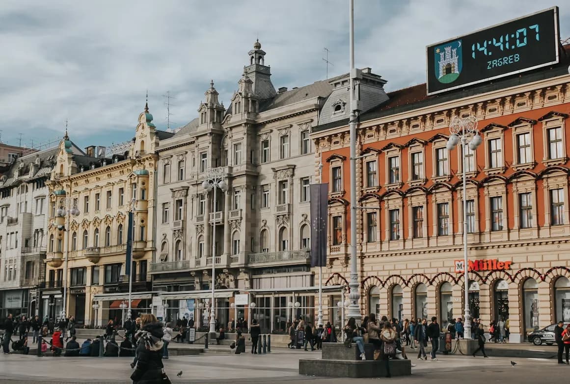 People gather in Ban Jelačić Square, the bustling heart of Zagreb, surrounded by beautiful historic buildings and a large digital clock.