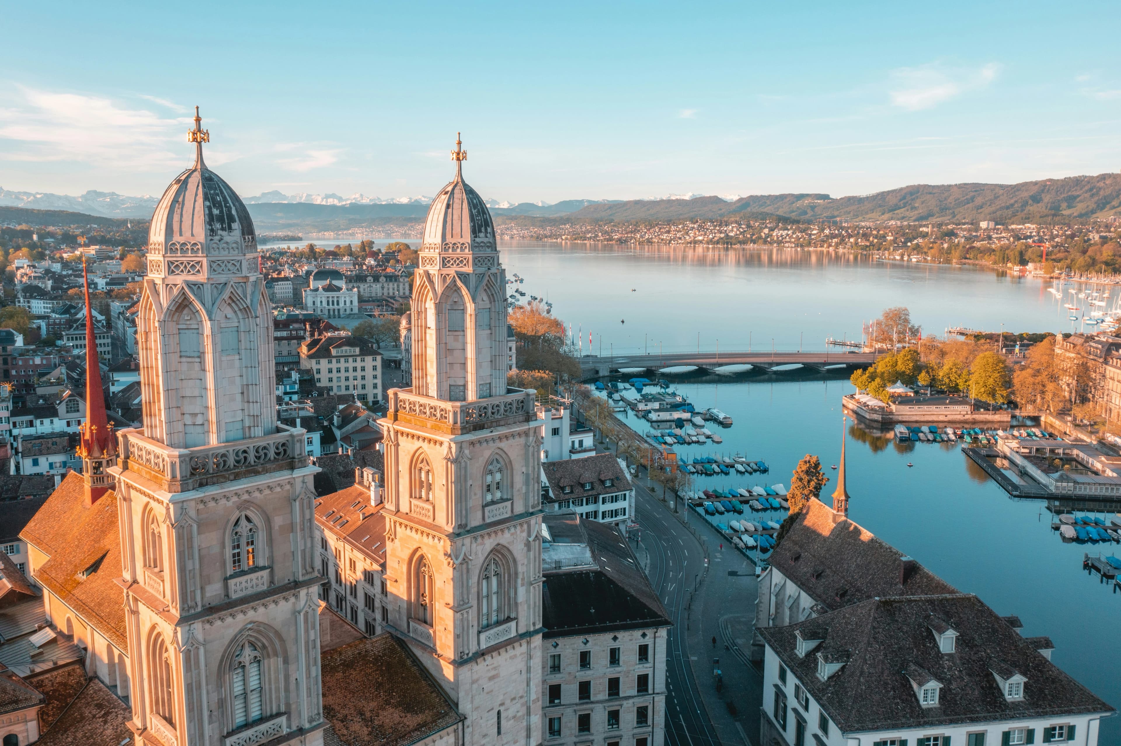An aerial view of the Grossmünster church towers overlooking Lake Zurich, with snow-capped mountains in the distance.
